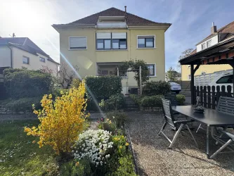 Ein schönes Mehrfamilienhaus mit einem gepflegten Garten. Im Vordergrund sind Blumen und eine Terrasse mit Stühlen und Tisch zu sehen.