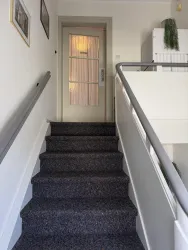 Eine Treppe mit dunklem Teppich führt zu einer Tür im Hintergrund. Der Raum ist hell und schlicht eingerichtet.