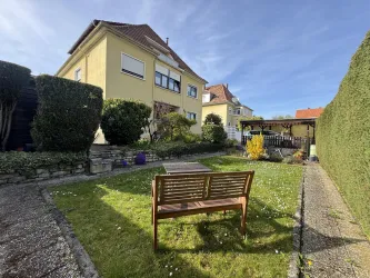 Ein ruhiger Garten mit einer Bank, umgeben von Sträuchern und Blumen. Im Hintergrund steht ein Ferienwohnung Hannelore unter blauem Himmel.