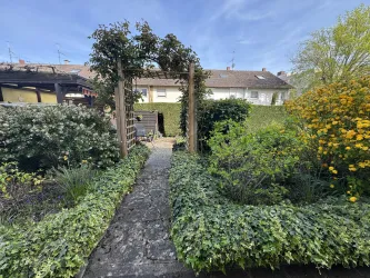 Ein schöner Gartenweg mit üppigem Grün und bunten Blumen. Im Hintergrund sind ein hübsches Haus und ein blauer Himmel sichtbar.