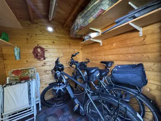 Ein kleiner Abstellraum aus Holz mit zwei Fahrrädern und ein paar Regalen. Im Hintergrund sind einige Sitzgelegenheiten und eine Lampe sichtbar.