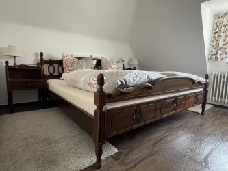 Ein gemütliches Schlafzimmer mit einem großen, klassischen Holzbett und einem Nachttisch. Die Wände sind grau und das Licht wirkt warm und einladend.