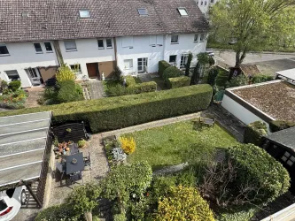 Ein gemütlicher Garten mit grünen Hecken und bunten Pflanzen. Im Hintergrund sind Reihenhäuser und eine asphaltierte Straße zu sehen.