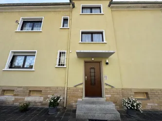 Ein gelbes Gebäude mit einem Eingang und mehreren Fenstern. Vor dem Eingang stehen Blumentöpfe mit weißen Blumen.