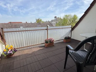 Ein gemütlicher Balkon mit einem Tisch und einem Stuhl. Blumentöpfe mit bunten Blumen schmücken die Ecke und die Aussicht zeigt ein ruhiges Stadtgebiet.