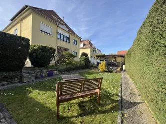Ein schöner Garten mit einer Holzbank, Hecken und blühenden Pflanzen. Im Hintergrund ist ein Gebäude und eine überdachte Sitzecke zu sehen.