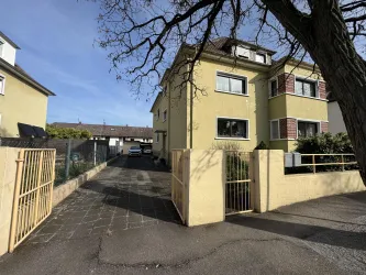 Ein gelbes Mehrfamilienhaus an einer ruhigen Straße. Der Eingang ist durch ein Tor erreichbar, und im Hintergrund sind weitere Gebäude zu sehen.