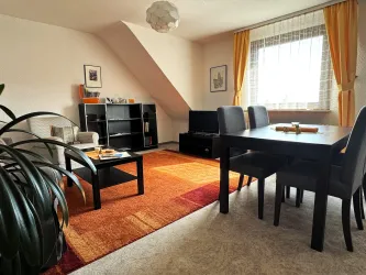 Ein gemütliches Wohnzimmer mit einem Essbereich und einem großen Teppich. Helle Vorhänge lassen viel Tageslicht herein.