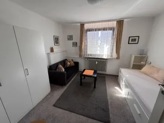 Ein einfaches Zimmer mit einem Bett, einem Sofa und einem Tisch. Die Fenster lassen viel Licht herein und die Wände sind hell gestrichen.