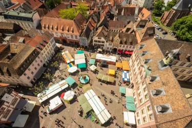 Ein belebter Marktplatz mit bunten Standen und Menschen. Die umliegenden Gebäude haben traditionelle, historische Architektur.