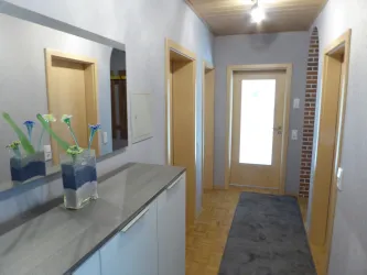 Ein heller Flur mit grauem Bodenbelag und mehreren Türen. Auf einem Sideboard steht eine Vase mit bunten Blumen.