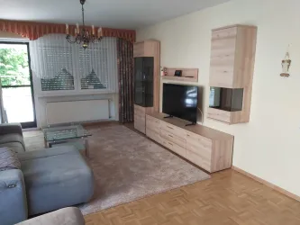 Ein modernes Wohnzimmer mit einem bequemen Sofa und einem gläsernen Couchtisch. Im Hintergrund steht eine Fernseherkonsole und große Fenster sorgen für viel Licht.