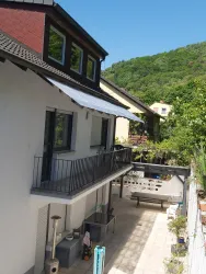 Ein modernes Haus mit einem Balkon und einem großen Innenhof. Im Hintergrund sind grüne Hügel und angrenzende Gebäude sichtbar.