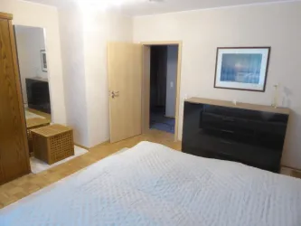 Ein helles Schlafzimmer mit einem großen Bett, einer Kommode und einem Spiegel. Die Wände sind neutral, und es gibt eine offene Tür zu einem weiteren Raum.