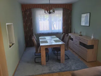 Ein gemütlicher Essbereich mit einem großen Holztisch und mehreren Stühlen. Das Fenster lässt viel Tageslicht herein, und die Wände sind in sanften Farben gestaltet.