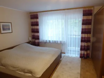 Ein Schlafzimmer mit einem großen Bett und einem Teppich. Vor dem Fenster hängen helle Vorhänge und es gibt einen Schrank an einer Wand.