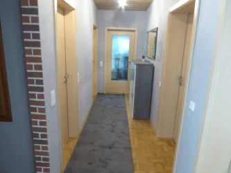 Ein heller Flur mit mehreren Türen und einem grauen Teppich. An einer Wand befindet sich eine schmale Kommode und ein Spiegel.