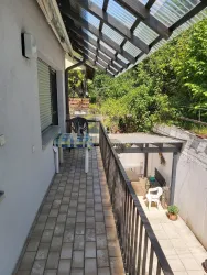 Ein schmaler Balkon mit Fliesen und einer Überdachung. Im Hintergrund sieht man einen grünen Garten und eine Terrasse mit Gartenmöbeln.