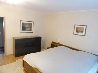 Ein gemütliches Schlafzimmer mit einem großen Bett und einem Holzschrank. An den Wänden hängen zwei Bilder, und der Raum hat einen neutralen Farbton.