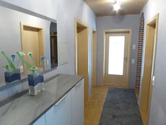 Ein heller Flur mit grauem Bodenbelag und einer großen Eingangstür. Auf der linken Seite steht ein moderner Schrank mit einer Vase und frischen Blumen.