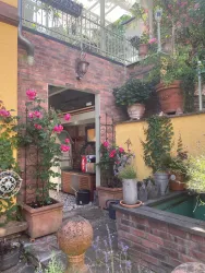 Ein schöner Innenhof mit roten Rosen und vielen Pflanzen in Töpfen. Im Hintergrund ist eine offene Tür, die durch das offene Gartenhaus zur Terrasse hinterm Haus führt.