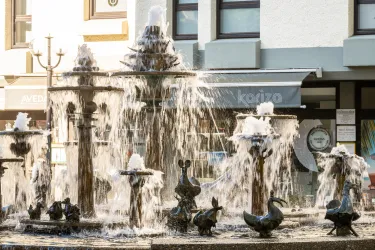 Ein schöner Brunnen mit künstlerischen Figuren und sprudelndem Wasser. Im Hintergrund sind Geschäfte sichtbar.