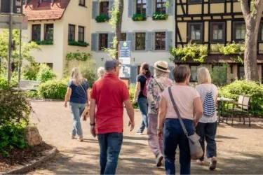 Eine Gruppe von Menschen spaziert durch eine malerische Straße mit traditionellen Häusern. Es ist sonnig und die Umgebung ist grün und einladend.