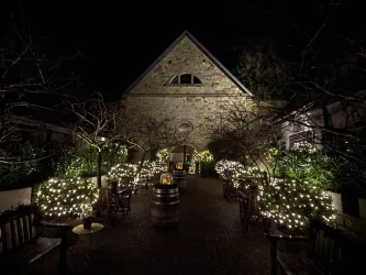 Eine stimmungsvolle, beleuchtete Gartenanlage bei Nacht mit Bäumen und Sitzgelegenheiten. Das Gebäude im Hintergrund hat eine charmante Steinfassade.