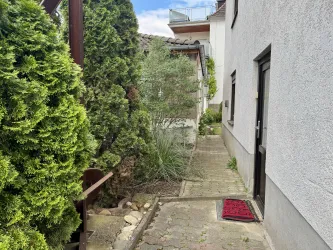 Ein schmaler Weg führt zwischen zwei Häusern vorbei, umgeben von grünen Sträuchern. Ein rotes Fußmatte liegt vor einer Wohnungstür.