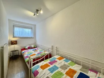 Ein helles Schlafzimmer mit zwei Einzelbetten und bunten Bettlaken. Durch ein Fenster scheint Tageslicht hinein.