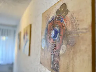 Ein Kunstwerk mit bunten Farben und abstrakten Formen hängt an der Wand. Im Hintergrund sind weitere Bilder zu sehen.