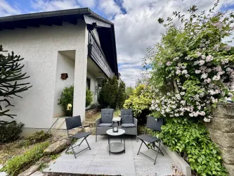 Eine gemütliche Sitzecke mit schwarzen Gartenstühlen und einem Tisch. Umgeben von üppigem Grün und blühenden Pflanzen.