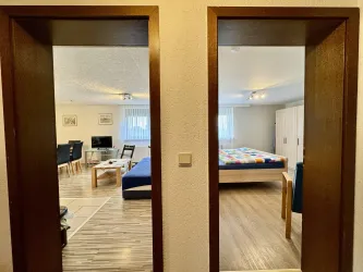 Zwei Türen, die auf zwei verschiedene Räume blicken. Der linke Raum hat eine gemütliche Sitzecke, während der rechte Raum ein Bett und einen Kleiderschrank zeigt.