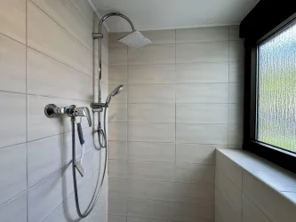 Eine moderne Duschkabine mit hellen Fliesen und einem großen Fenster. Die Dusche ist mit einem Handbrausekopf und einem festen Duschkopf ausgestattet.
