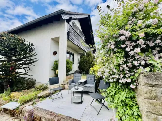 Ein gemütlicher Sitzbereich vor einem Haus, umgeben von grünen Pflanzen und blühenden Sträuchern. Die Terrasse bietet Platz für Entspannung im Freien.