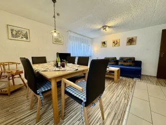 Ein gemütlicher Essbereich mit einem Holztisch und schwarzen Stühlen. Im Hintergrund ist ein Wohnzimmer mit einem blauen Sofa und bunten Bildern an den Wänden zu sehen.