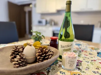Ein Tisch mit einer Flasche Bacchus-Wein, einer Kerze und einer Schale mit Kiefernzapfen und einem dekorativen Ei. Im Hintergrund ist eine helle Küche zu sehen.
