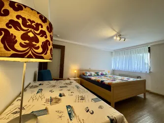 Ein helles Zimmer mit zwei Betten und einem großen Fenster. Ein dekorativer Lampenschirm sorgt für gemütliches Licht.