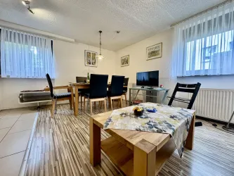 Ein gemütliches Wohnzimmer mit einem Esstisch und Stühlen. Es gibt einen Fernseher und große Fenster mit Vorhängen.