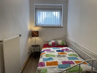 Ein kleines, helles Schlafzimmer mit einem Einzelbett und einem bunten Bettüberwurf. Die Wand ist weiß, und ein Fenster sorgt für Tageslicht.