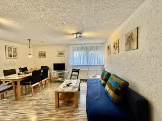 Eine gemütliche Wohnung mit einem blauen Sofa und einem Esstisch. Es gibt viele Fenster und dekorative Bilder an den Wänden.