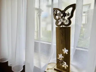 Eine dekorative Holzfigur in Form eines Schmetterlings steht auf einem Tisch. Vor einer hellen Fensterbank mit weißen Vorhängen sind kleine Blumenmuster zu sehen.