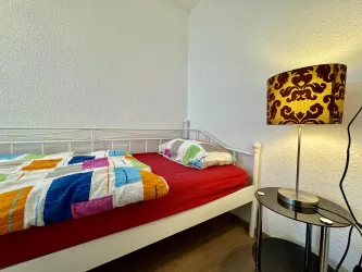 Ein einfaches Schlafzimmer mit einem weißen Bett und bunten Bettdecken. Neben dem Bett steht eine dekorative Lampe auf einem schwarzen Tisch.
