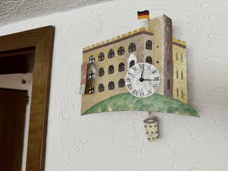 Eine dekorative Wanduhr in Form eines Schlosses mit einer deutschen Flagge. Sie hat ein rundes Zifferblatt und ist an einer weißen Wand befestigt.