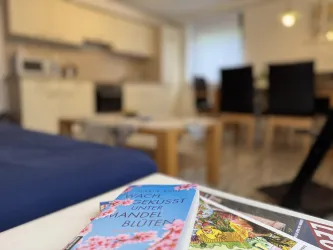 Ein gemütlicher Wohnraum mit einer modernen Küche im Hintergrund. Auf einem Tisch liegen Zeitschriften und ein Buch mit dem Titel „Wachgeküsst unter Mandelblüten“.