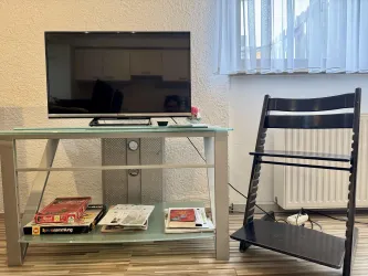 Ein Wohnzimmer mit einem Fernseher auf einem modernen Tisch. Darunter liegen Zeitschriften und daneben steht ein schwarzer Stuhl.