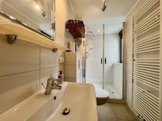 Ein modernes Badezimmer mit einem Waschbecken und einer ebenerdigen Dusche. Die Wände sind hell gefliest und es gibt viel Tageslicht.