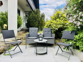 Eine gemütliche Terrasse mit zwei grauen Stühlen und einem Tisch in der Mitte. Umgeben von grünen Pflanzen und Blumen.