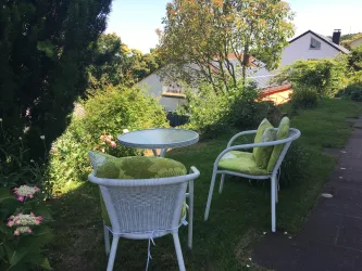 Ein gemütlicher Gartensitzbereich mit zwei weißen Stühlen und einem runden Tisch. Umgeben von üppigem Grün und Blumen bietet dieser Ort eine einladende Atmosphäre zum Entspannen.