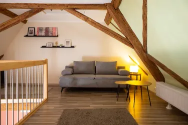Ein gemütlicher Wohnbereich unter dem Dach mit Holzbalken. Das Zimmer enthält ein graues Sofa, einen runden Tisch und eine Lampe.